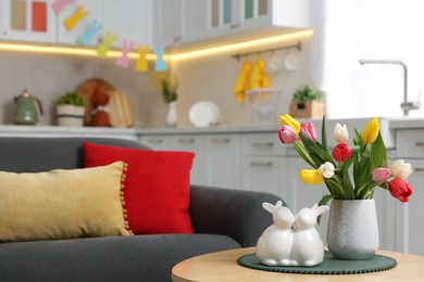 Easter decorations. Bouquet of tulips and bunny figures on table near sofa indoors. Space for text