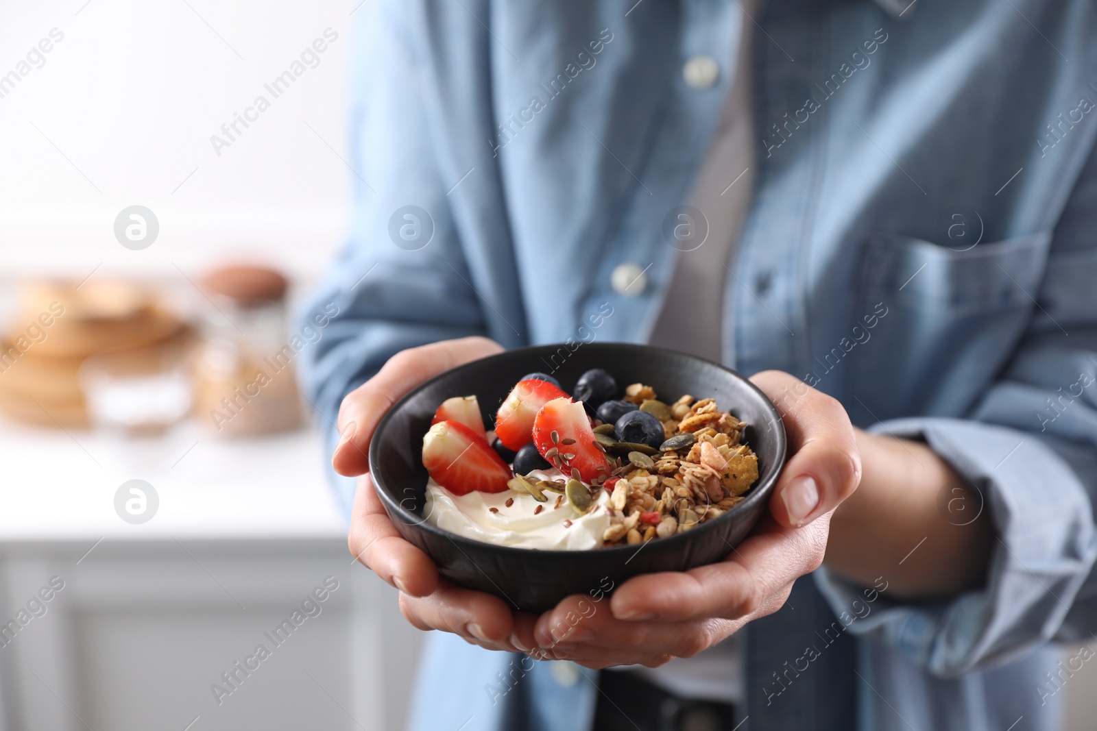 Photo of Woman holding bowl of tasty granola with berries, yogurt and seeds indoors, closeup