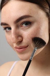 Beautiful woman with freckles applying makeup with brush on grey background, closeup