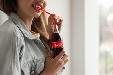 MYKOLAIV, UKRAINE - NOVEMBER 28, 2018: Young woman with bottle of Coca-Cola indoors, closeup. Space for text