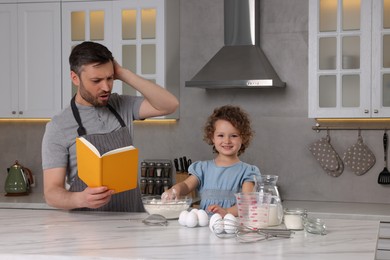 Photo of Cute little girl and her father with recipe book cooking in kitchen
