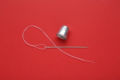 Silver thimble, needle and thread on red background, flat lay