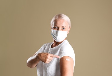 Mature woman in protective mask pointing at arm with bandage after vaccination on beige background