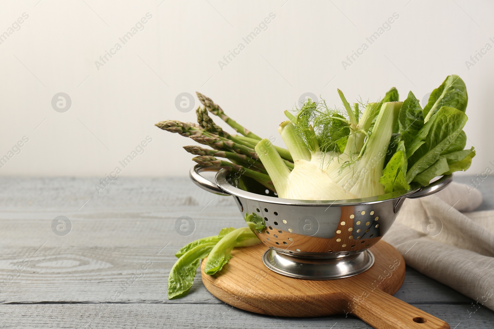 Photo of Metal colander with fennel, lettuce and asparagus on gray wooden table, space for text