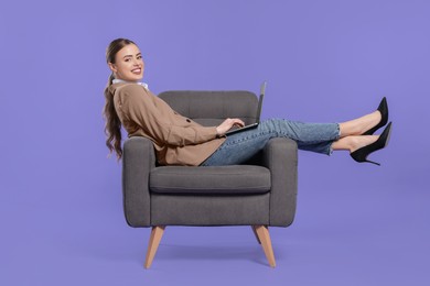 Happy woman with laptop sitting in armchair on violet background