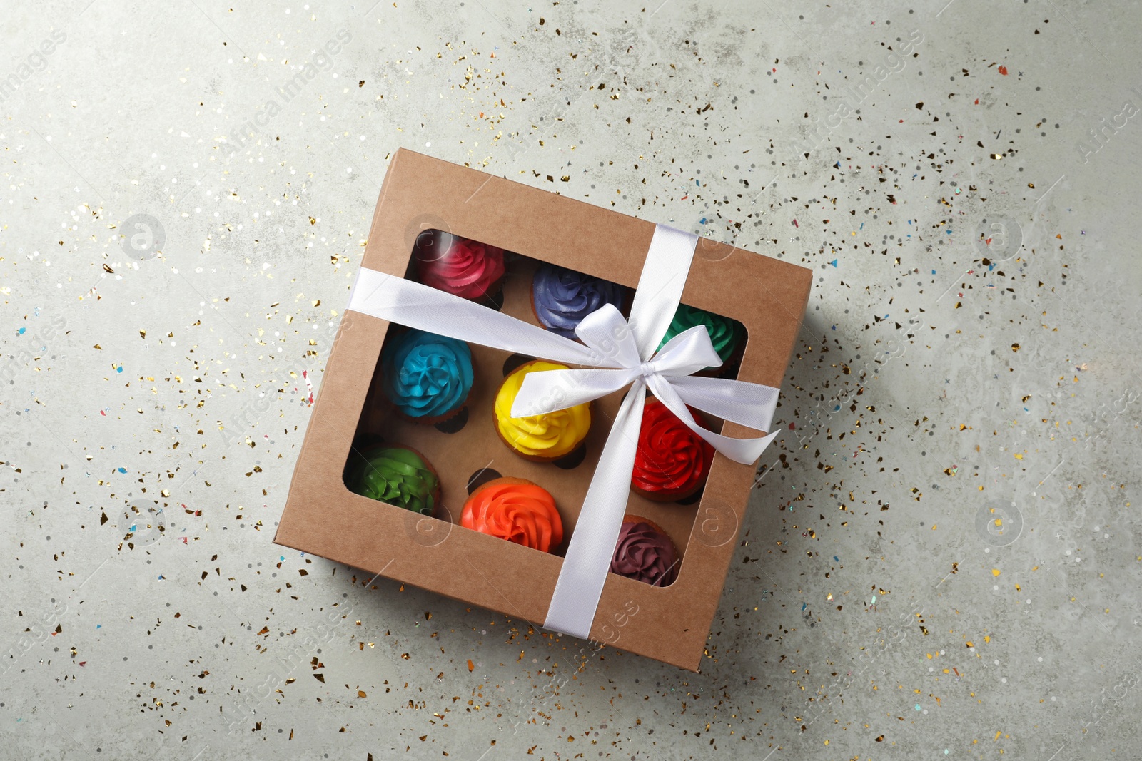 Photo of Box with delicious colorful cupcakes and confetti on light grey table, flat lay