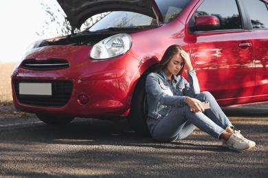 Stressed woman with smartphone near broken car outdoors