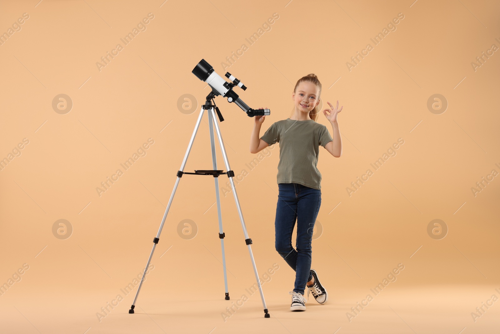 Photo of Happy little girl with telescope showing ok gesture on beige background