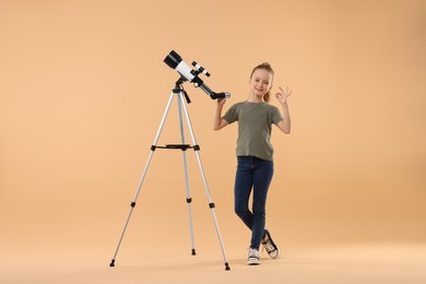 Happy little girl with telescope showing ok gesture on beige background