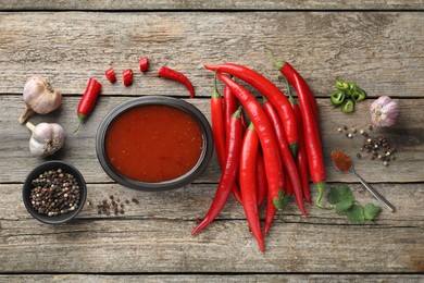 Photo of Spicy chili sauce in bowl and ingredients on wooden table, flat lay