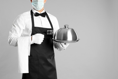 Photo of Waiter in medical face mask holding tray with lid on light grey background, closeup. Space for text