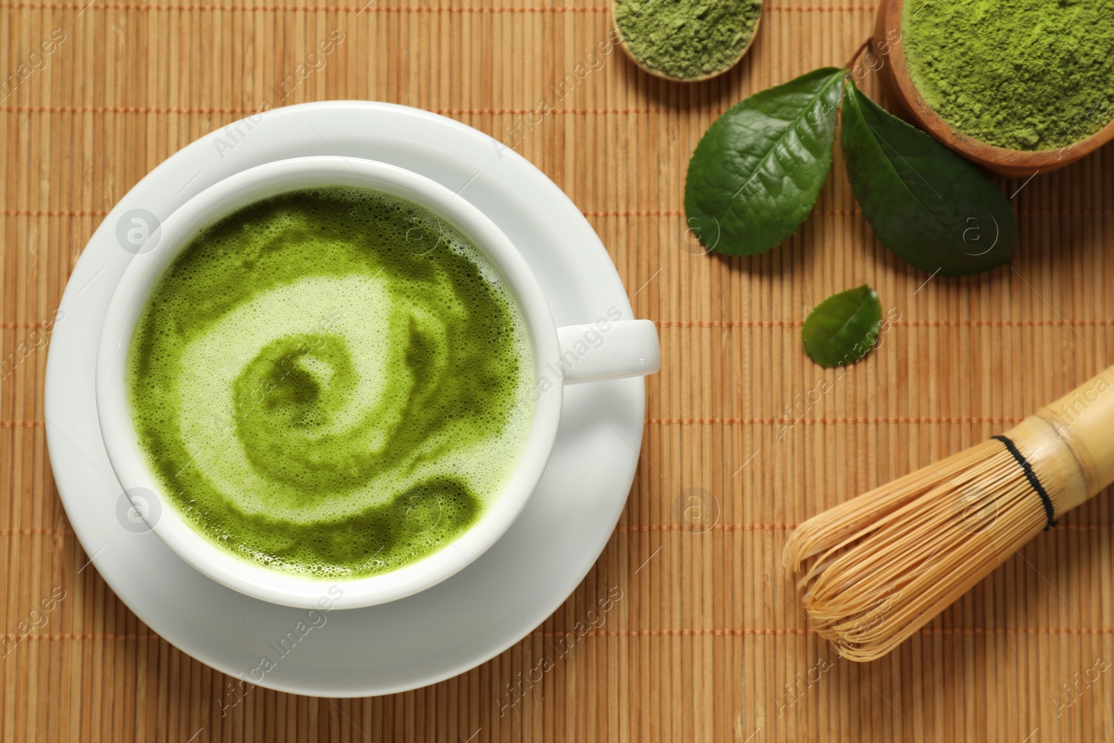 Photo of Delicious matcha latte in cup, powder, whisk and leaves on bamboo mat, flat lay