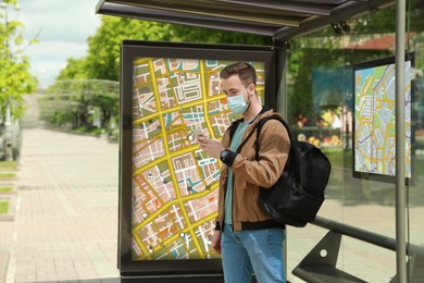 Image of Young man in protective mask with smartphone and backpack waiting for public transport at bus stop