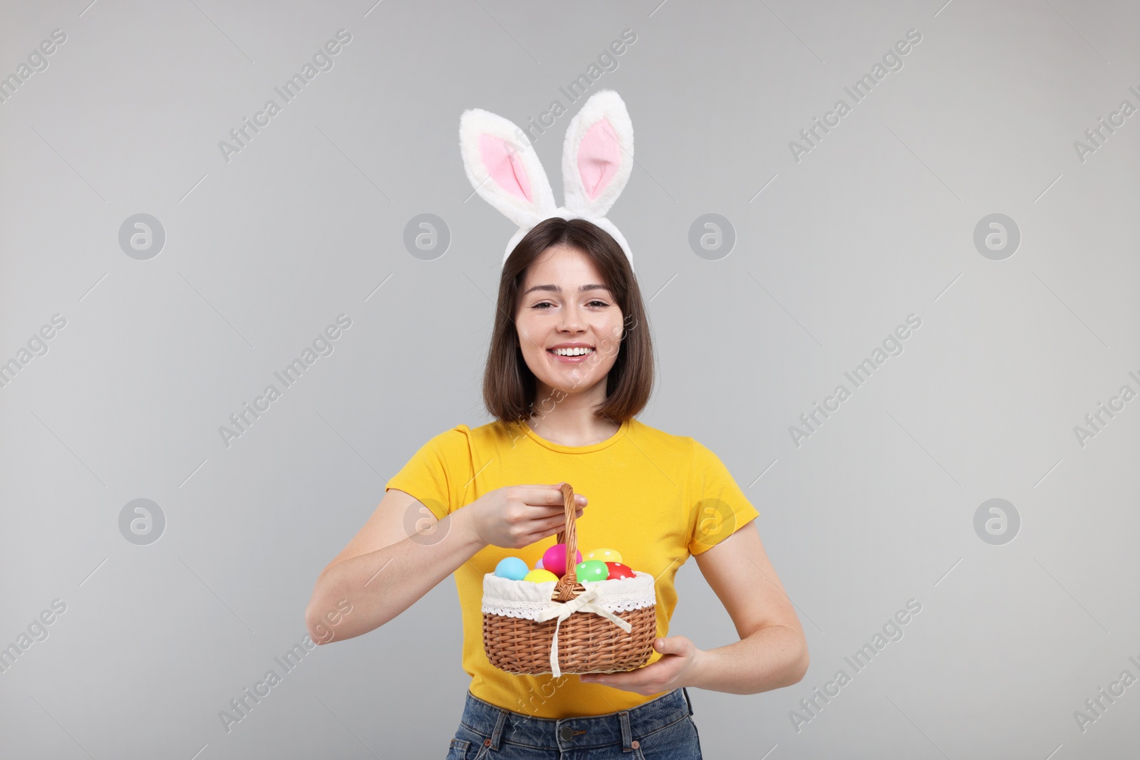 Photo of Easter celebration. Happy woman with bunny ears and wicker basket full of painted eggs on grey background