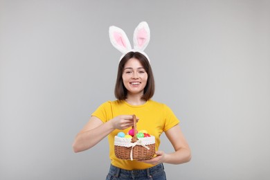 Photo of Easter celebration. Happy woman with bunny ears and wicker basket full of painted eggs on grey background