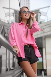 Photo of Beautiful young woman in stylish sunglasses near building outdoors