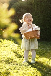 Happy little girl holding wicker basket with cute rabbit outdoors on sunny day