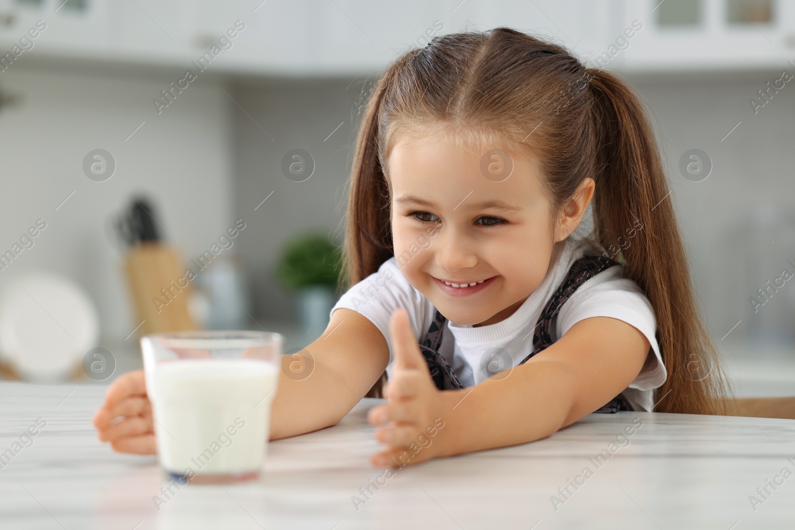 Photo of Cute girl reaching out for glass of milk at white table in kitchen