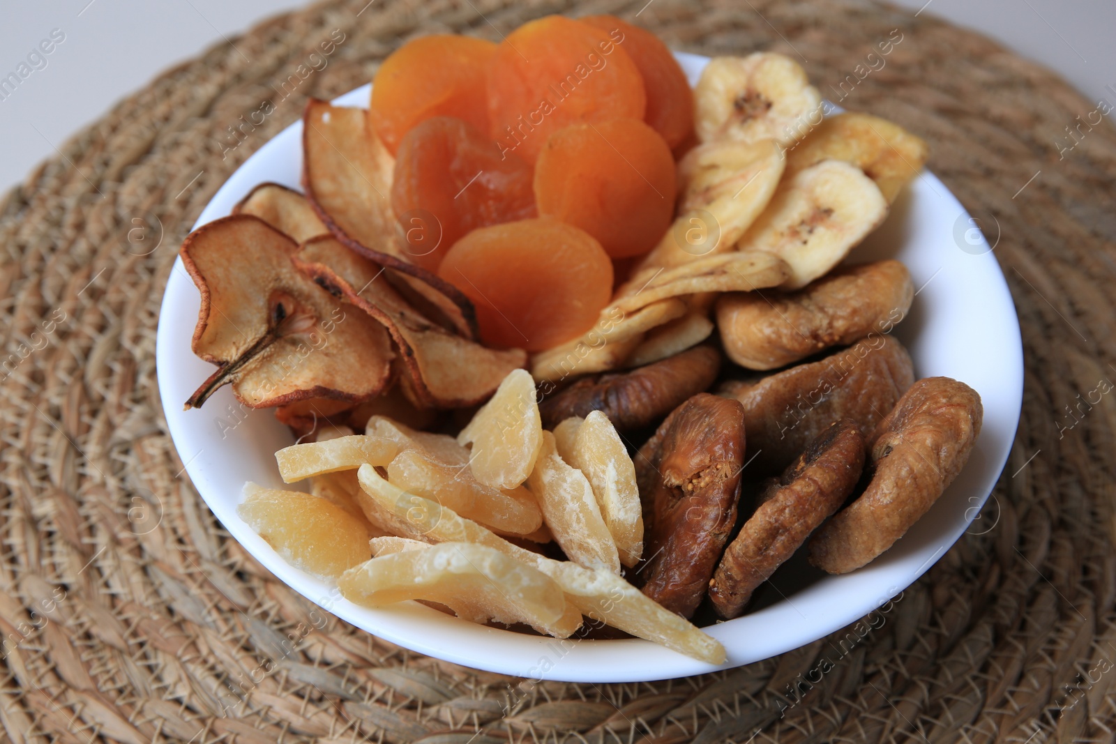 Photo of Bowl with different dried fruits on wicker mat, closeup