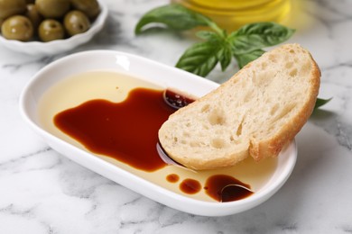 Photo of Bowl of organic balsamic vinegar with oil served with bread slice, basil and olives on white marble table, closeup