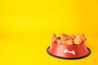 Bone shaped dog cookies in feeding bowl on yellow background, space for text
