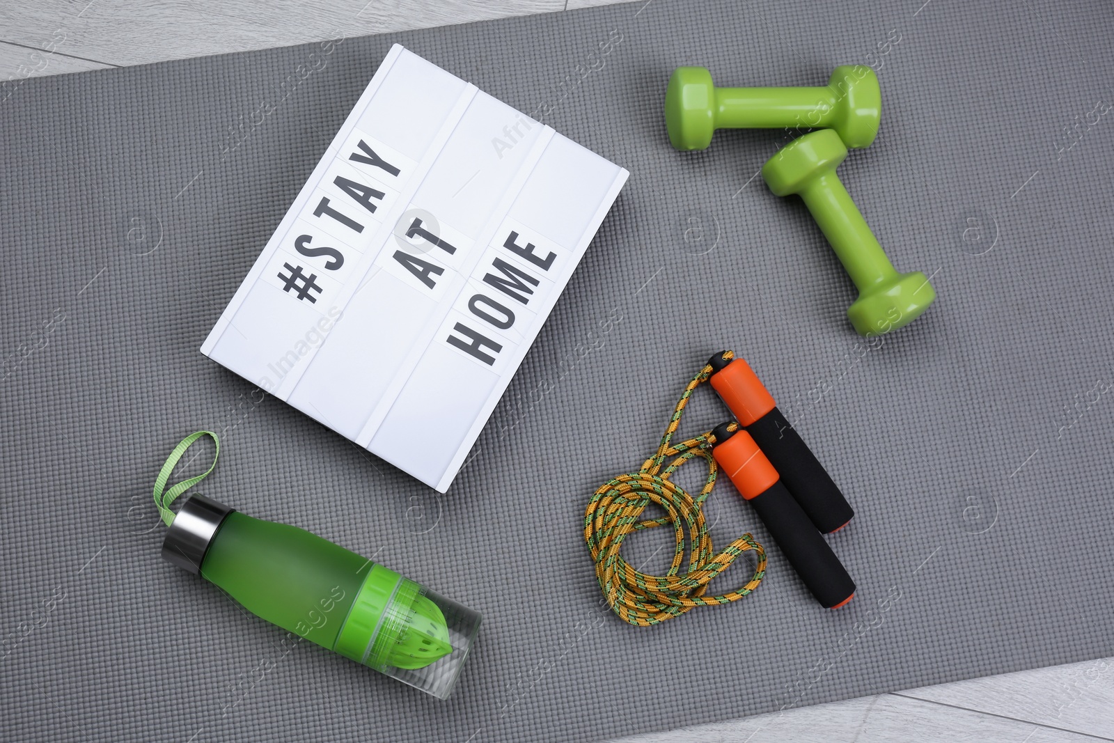 Photo of Sport equipment and lightbox with hashtag STAY AT HOME on grey yoga mat, flat lay. Message to promote self-isolation during COVID‑19 pandemic