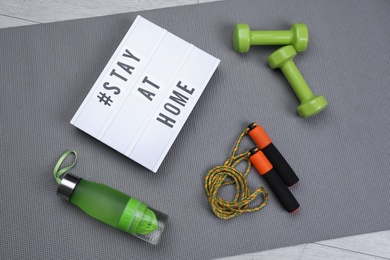 Sport equipment and lightbox with hashtag STAY AT HOME on grey yoga mat, flat lay. Message to promote self-isolation during COVID‑19 pandemic
