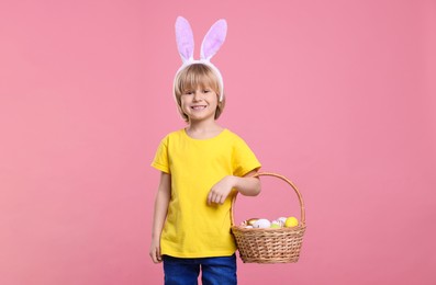 Photo of Happy boy in bunny ears headband holding wicker basket with painted Easter eggs on pink background