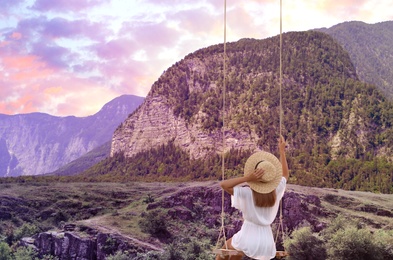 Image of Dream world. Young woman swinging over mountains under sunset sky 