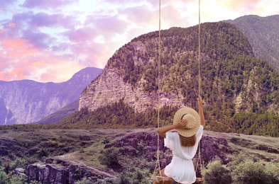 Dream world. Young woman swinging over mountains under sunset sky 