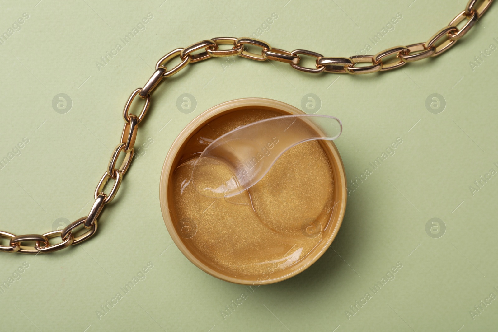 Photo of Under eye patches in jar with spatula and necklace on olive background, flat lay. Cosmetic product