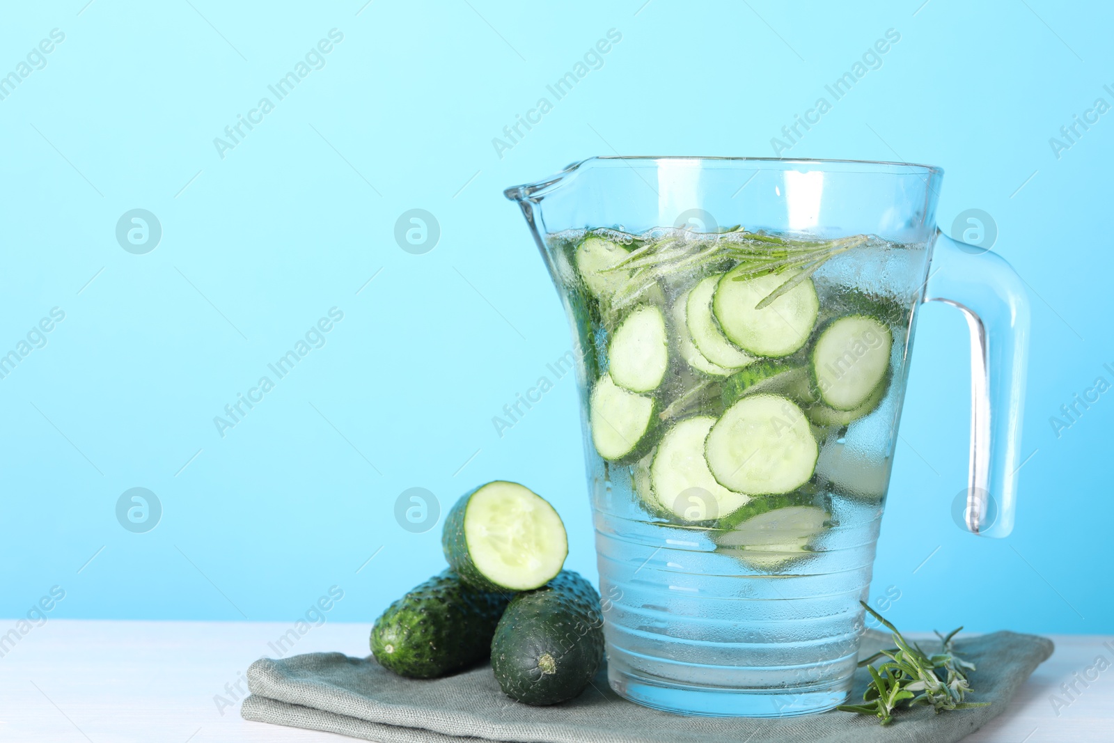 Photo of Refreshing cucumber water with rosemary in jug and vegetables on white table against light blue background. Space for text