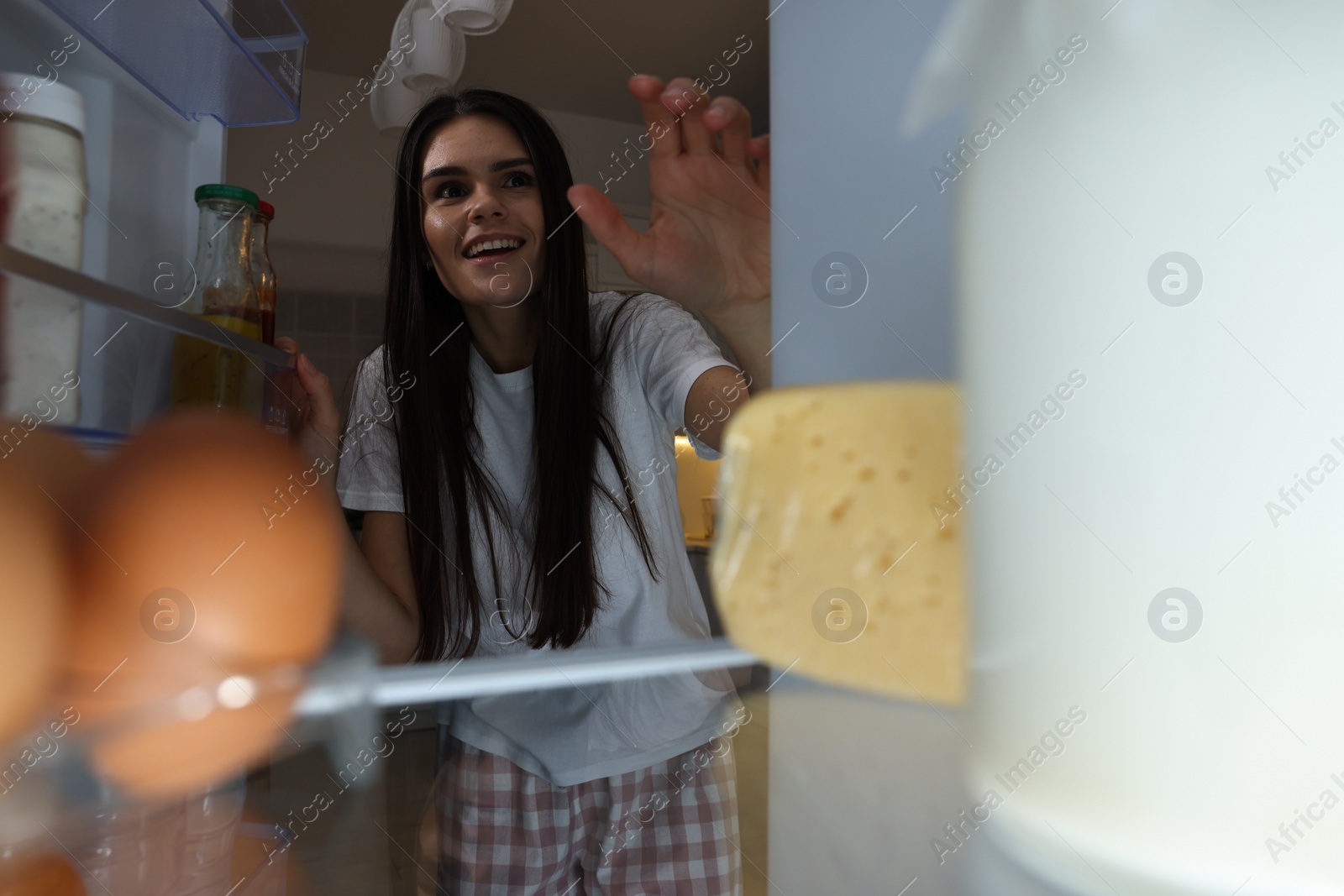 Photo of Young woman near modern refrigerator in kitchen at night, view from inside