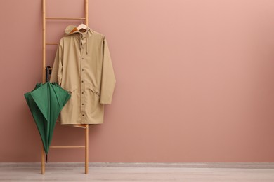 Photo of Stylish green umbrella and raincoat on wooden ladder near beige wall indoors. Space for text