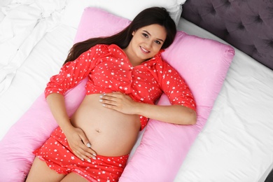 Beautiful pregnant woman lying with maternity pillow on bed at home, top view