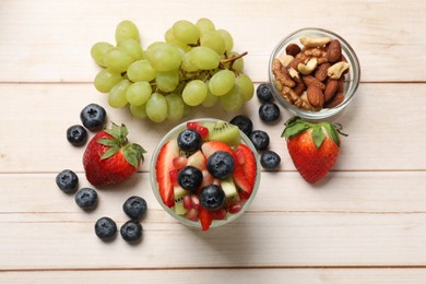 Photo of Healthy breakfast. Delicious fruit salad in glass, nuts and ingredients on light wooden table, flat lay