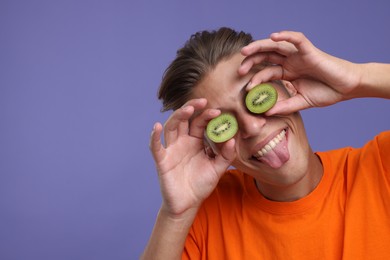 Photo of Smiling man covering eyes with halves of kiwi and showing tongue on violet background, closeup. Space for text