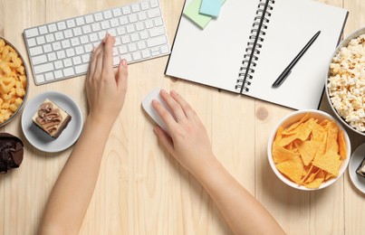 Bad eating habits. Woman working on computer at wooden table with different snacks, top view