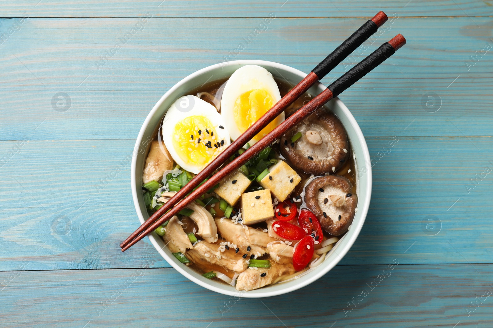 Photo of Bowl of delicious ramen and chopsticks on light blue wooden table, top view. Noodle soup