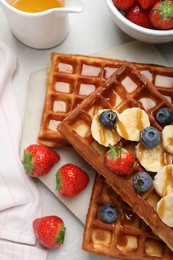 Photo of Board with delicious Belgian waffles, banana, berries and caramel sauce on light table, flat lay