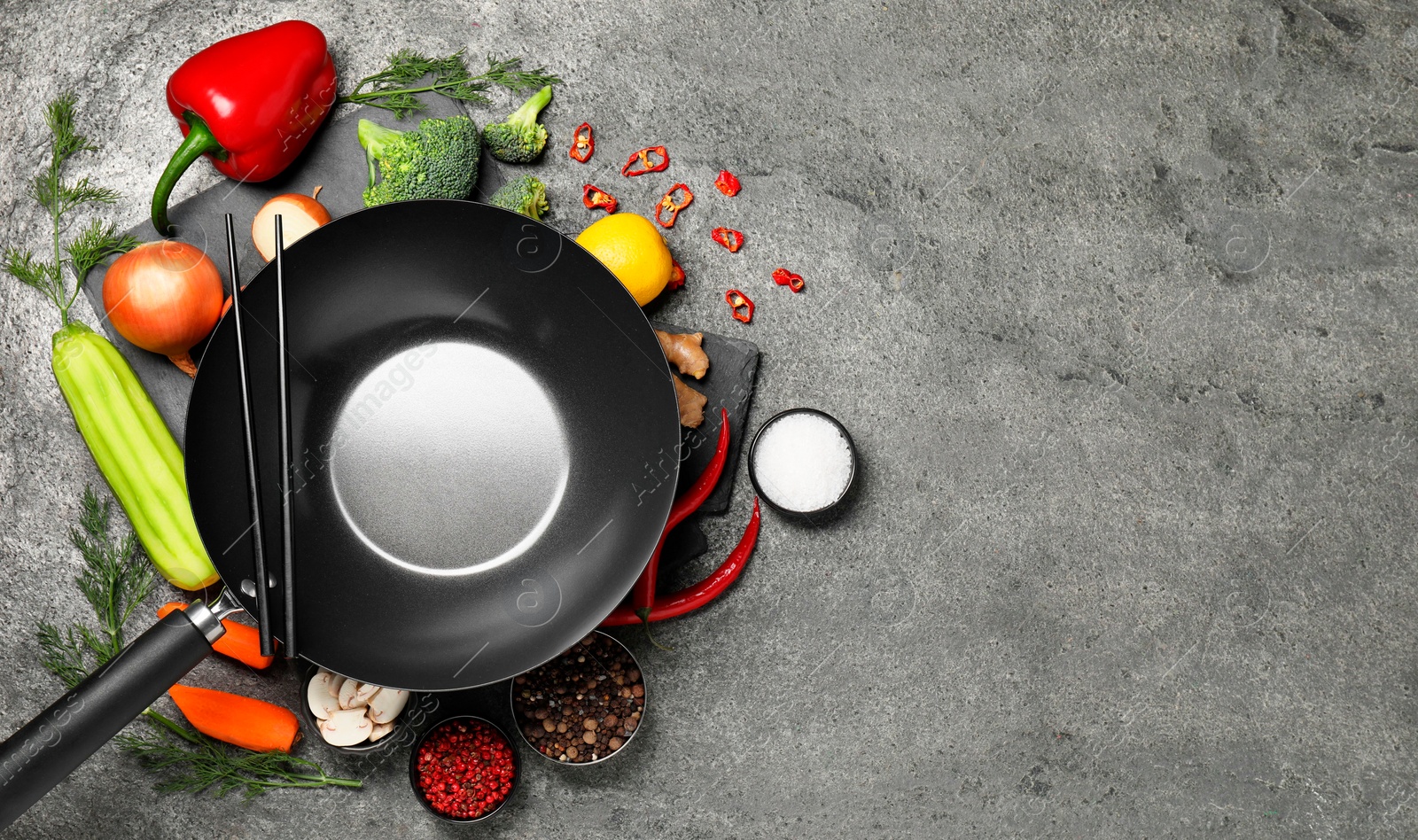 Photo of Empty iron wok and chopsticks surrounded by ingredients on grey table, flat lay. Space for text