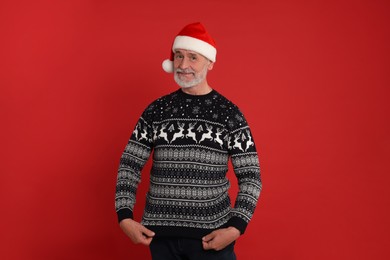 Photo of Senior man in Santa hat showing his Christmas sweater on red background