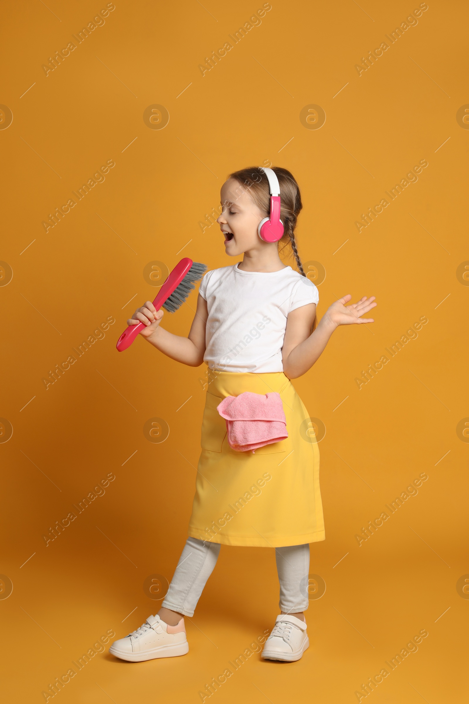 Photo of Cute little girl in headphones with brush singing on orange background