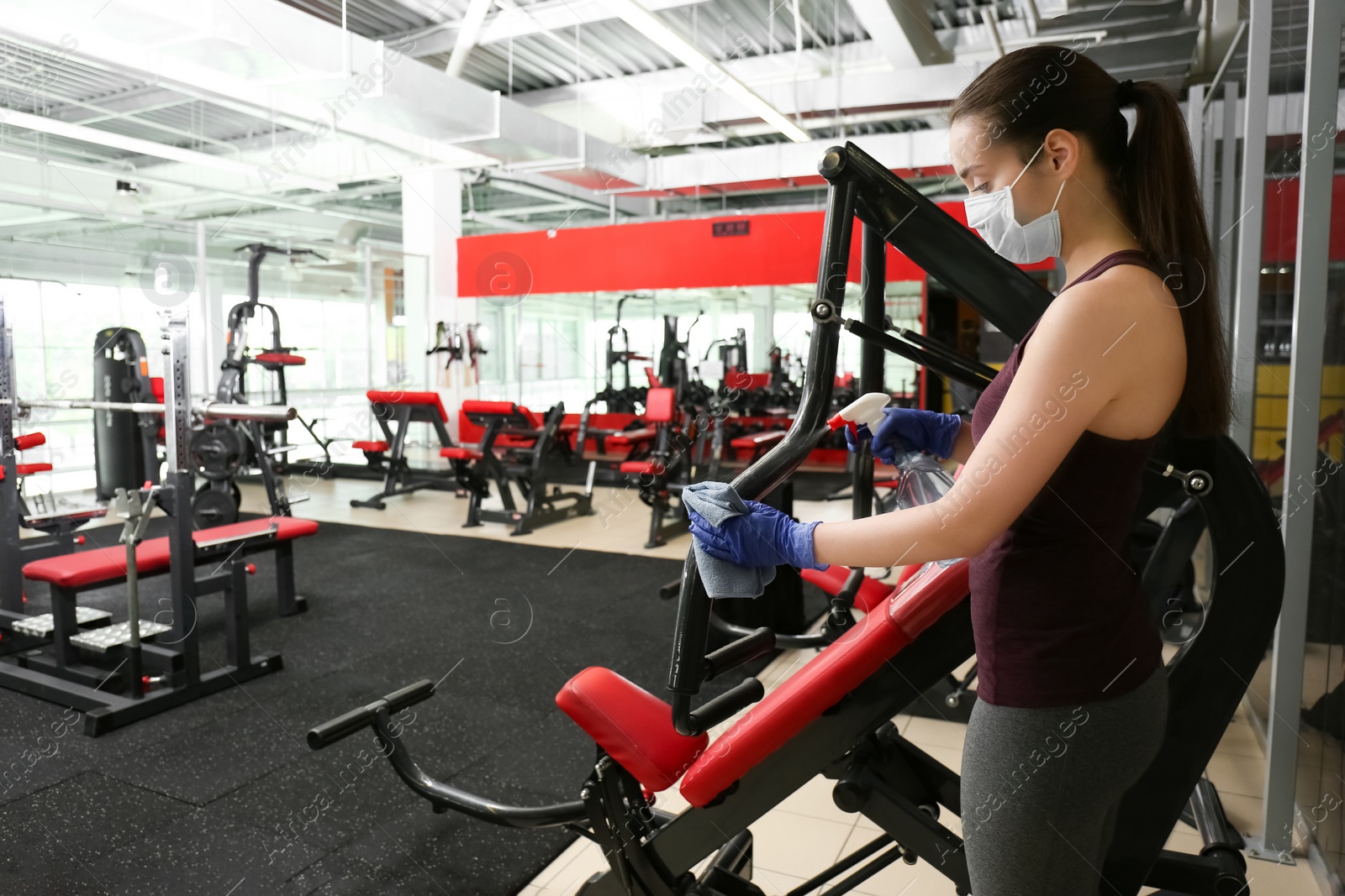 Photo of Woman cleaning exercise equipment with disinfectant spray and cloth in gym