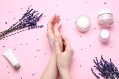 Woman applying hand cream and lavender flowers on pink background, top view