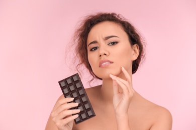 Beautiful young woman with acne problem holding chocolate on color background. Skin allergy