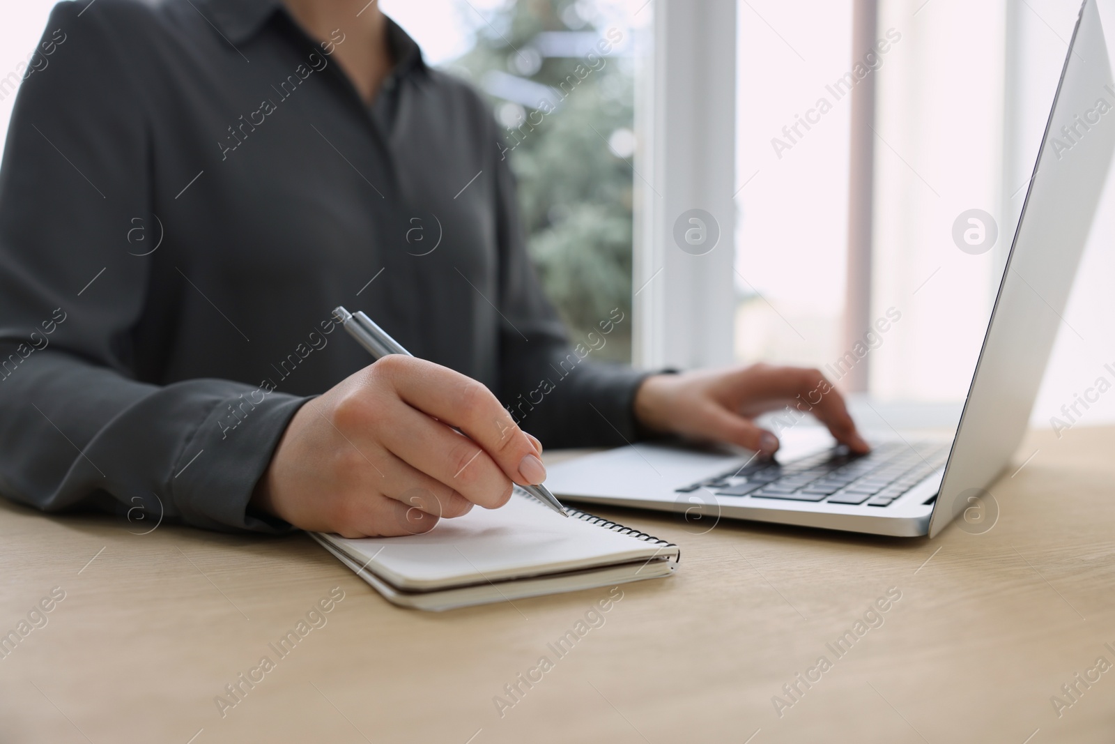 Photo of Woman with notebook and pen working on laptop at wooden table, closeup. Electronic document management