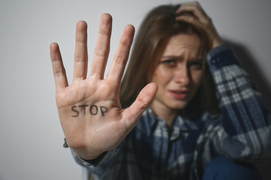 Crying young woman showing palm with word STOP near white wall, focus on hand. Domestic violence concept