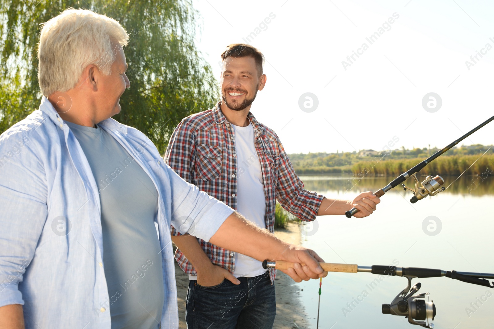 Photo of Father and adult son fishing together from riverside on sunny day