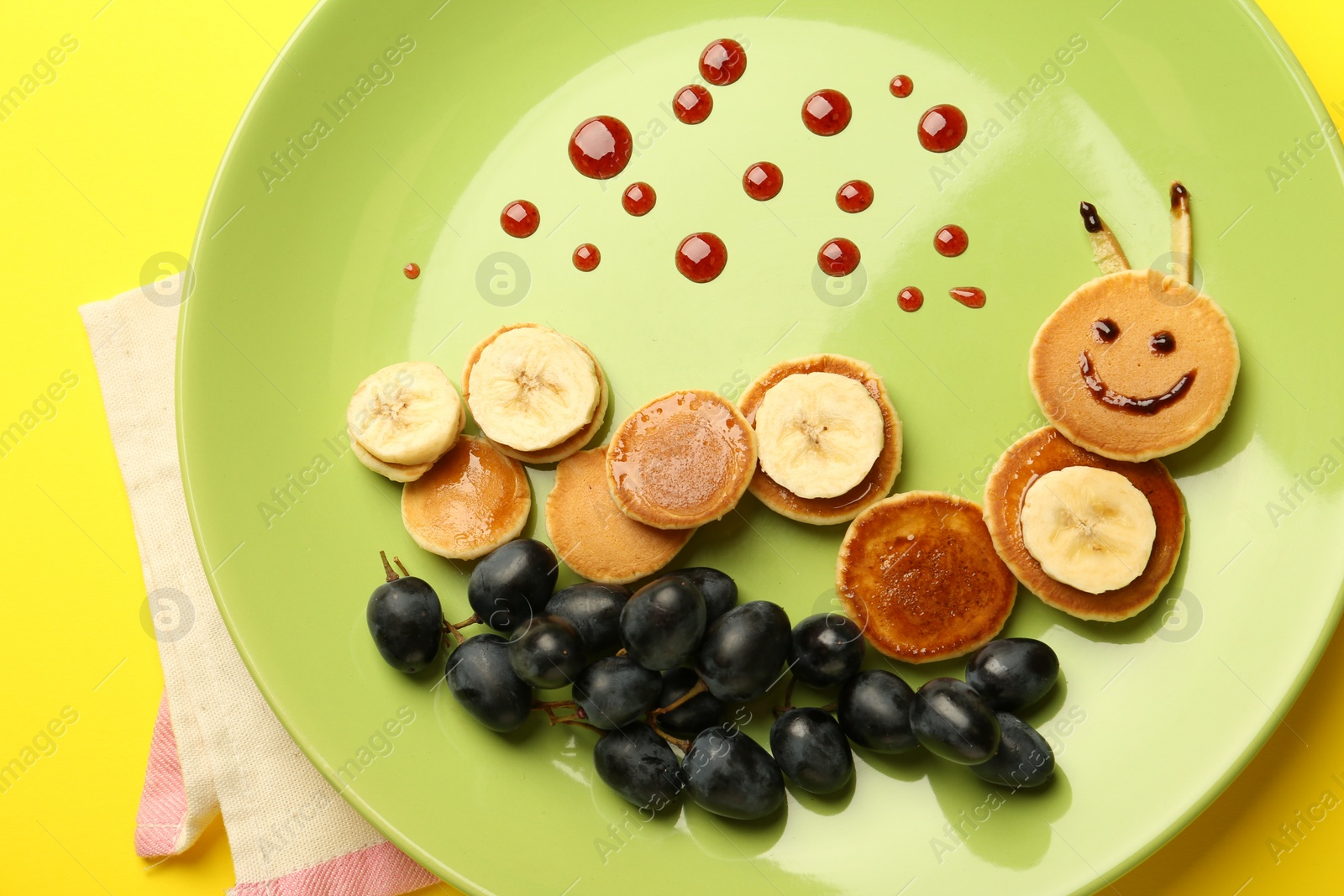 Photo of Creative serving for kids. Plate with cute caterpillar made of pancakes, grapes and banana on yellow background, top view